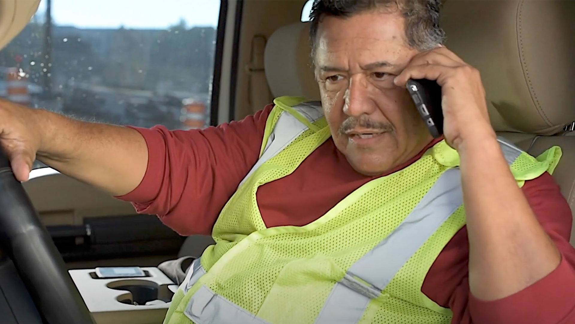Older male driver with right hand on the wheel talks holding cellphone to his left ear. 